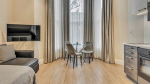 A modern living room with a flat-screen TV, a sofa, a glass dining table with chairs, and a kitchen area with a stove. - Cleveland Residences Chelsea (London)