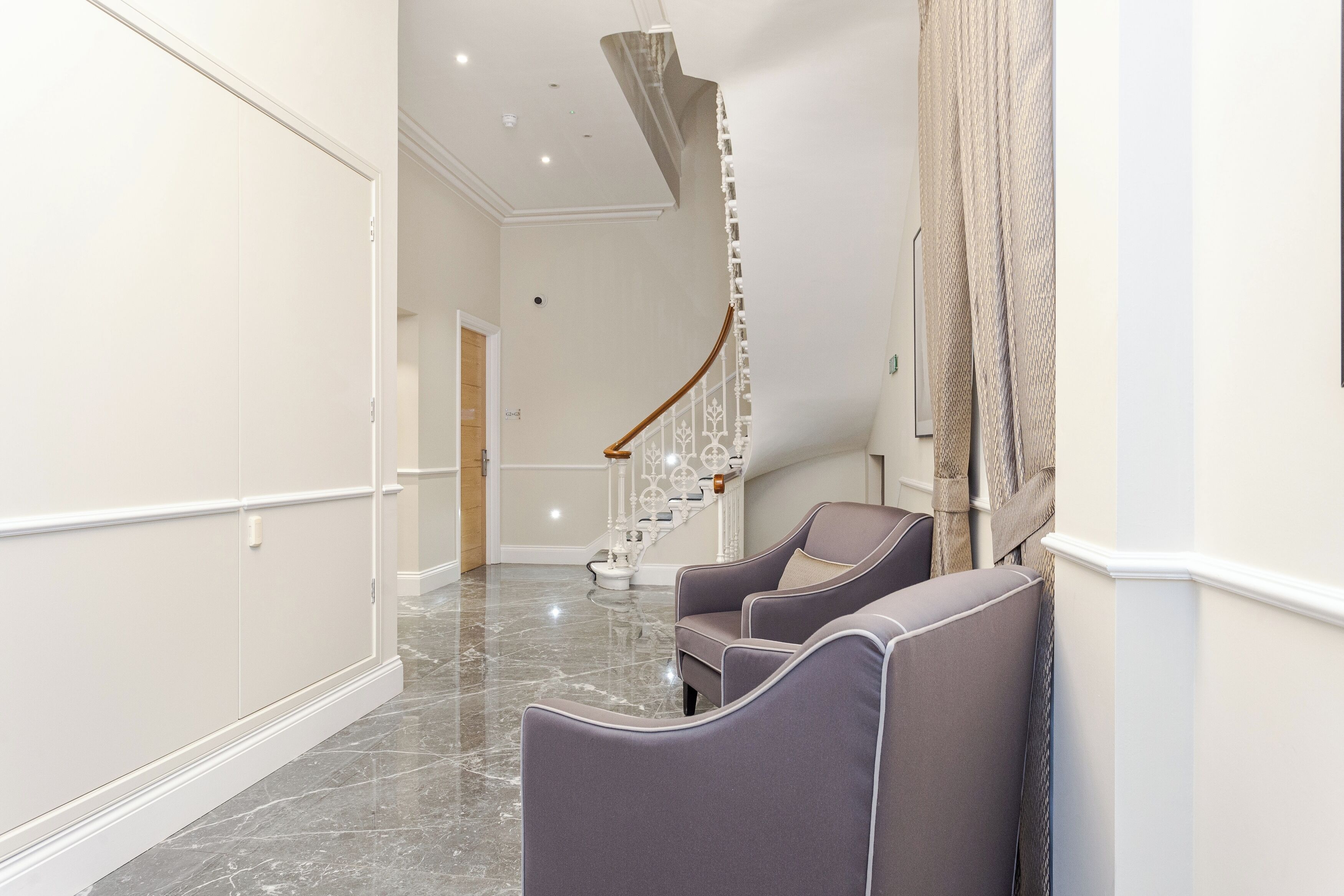 A spacious hallway with a marble floor, a staircase with a wooden handrail, and two grey armchairs.