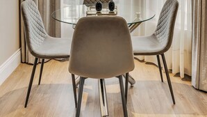 A dining area with a glass table and two chairs, a small decorative object on the table, and a view of buildings through sheer curtains. - Cleveland Residences Chelsea (London)