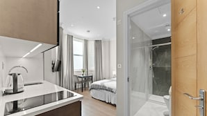 A modern hotel room with a kitchen area, a bed, a shower, and a glass door leading to a bathroom. - Cleveland Residences Chelsea (London)
