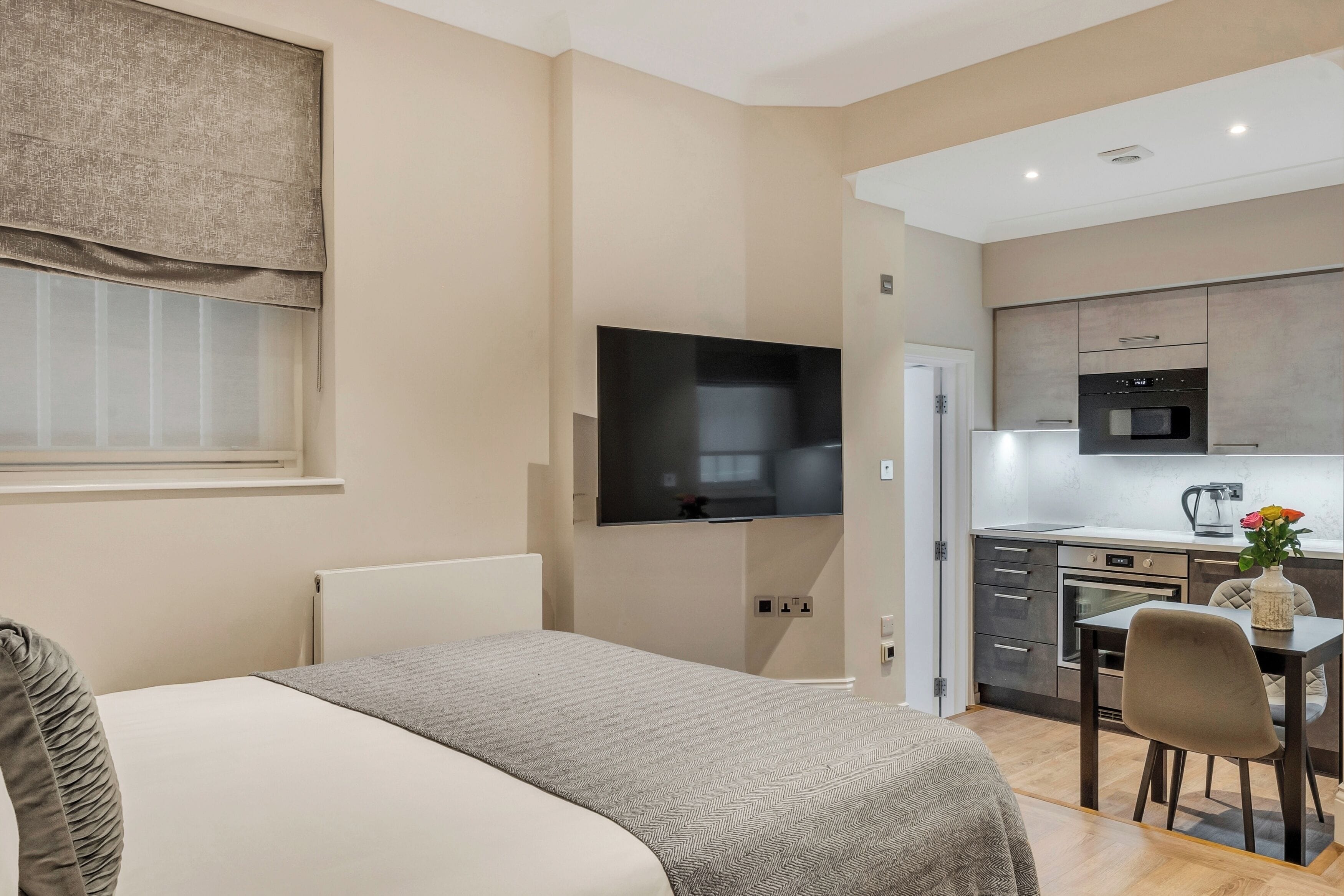A modern hotel room with a bed, a TV mounted on the wall, a kitchenette with a microwave, oven, and sink, and a small dining area with a table and chairs.