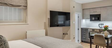 A modern hotel room with a bed, a TV mounted on the wall, a kitchenette with a microwave, oven, and sink, and a small dining area with a table and chairs.
