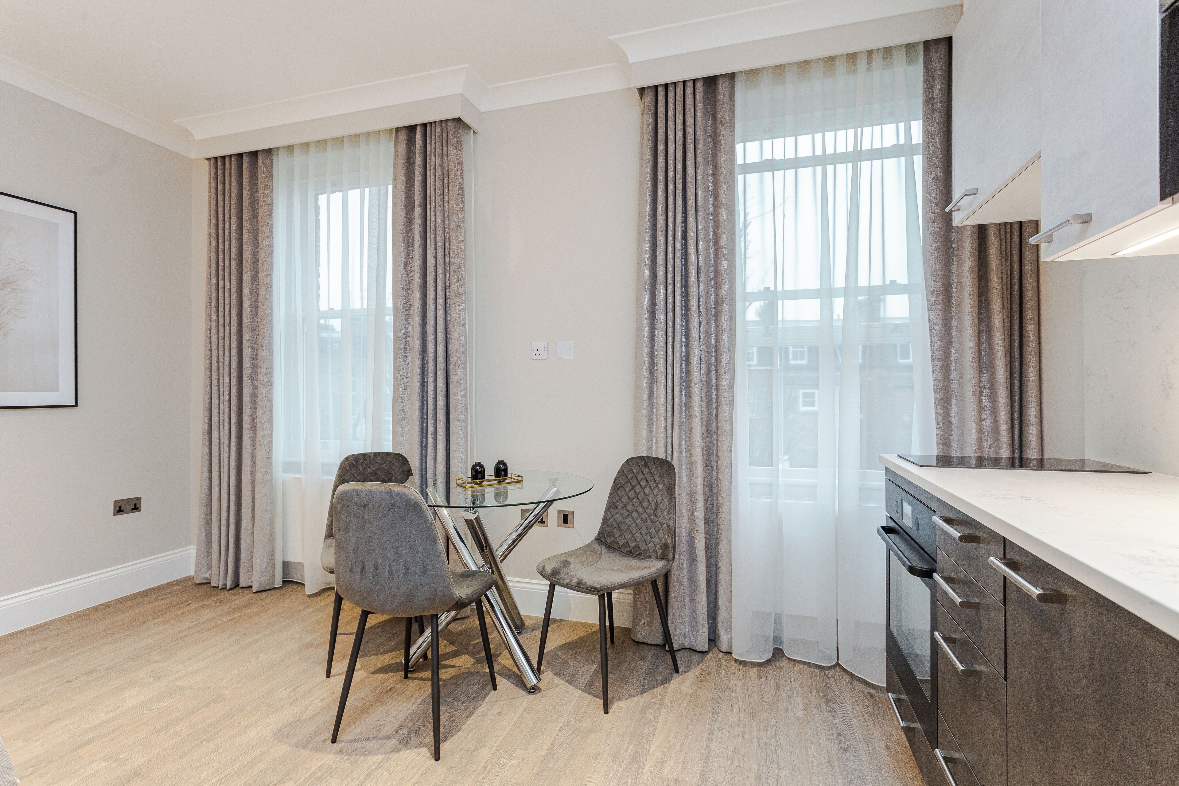 A modern kitchen with a built-in oven, a dining area with two chairs and a glass table, and large windows with curtains.