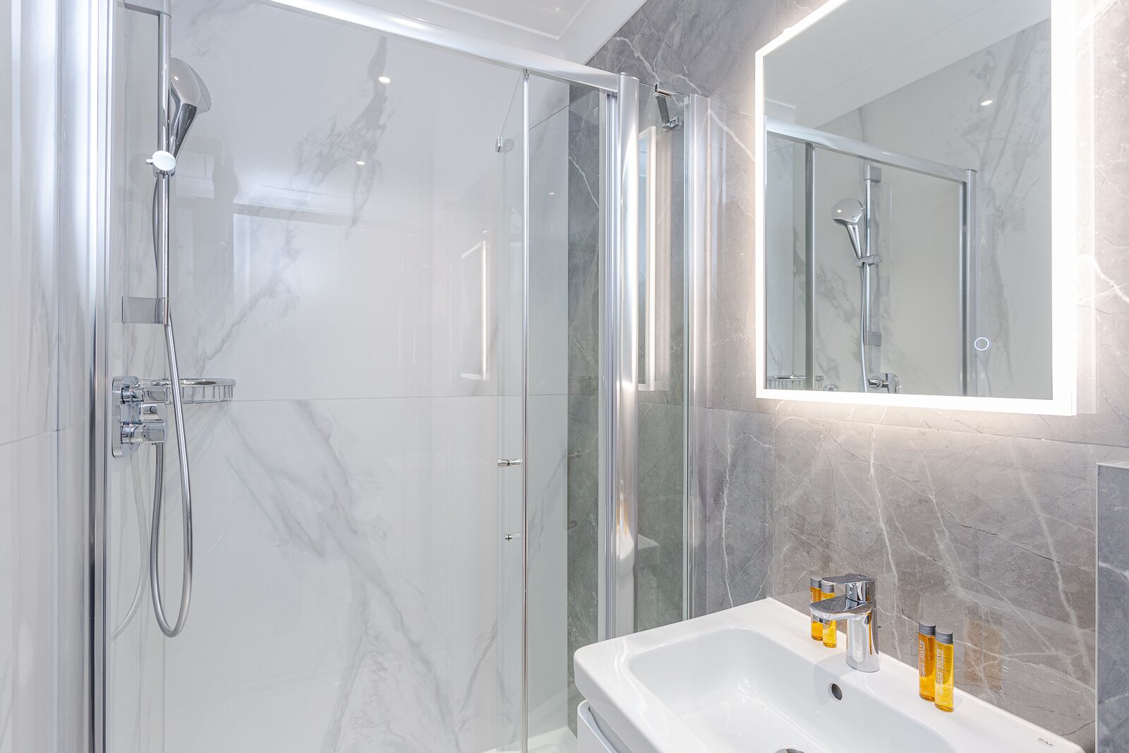 A modern bathroom with a glass-enclosed shower, a white sink with two bottles of shampoo, and a large mirror above the sink.