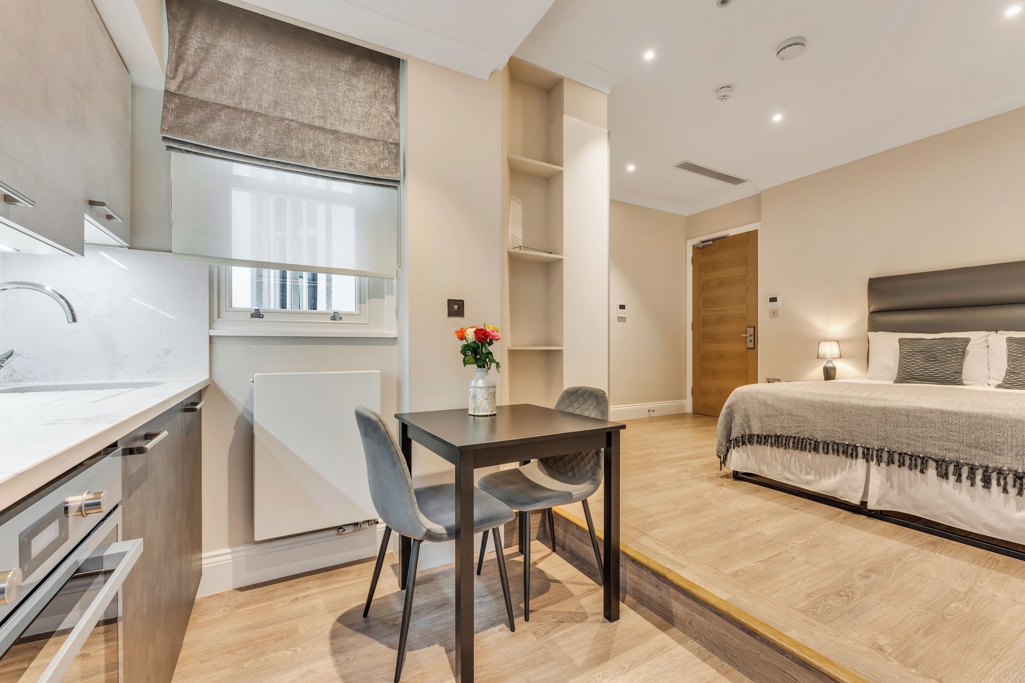 Una habitación de hotel moderna con cocina, una mesa de comedor con dos sillas, una cama con cabecero y una mesita de noche con lámpara.