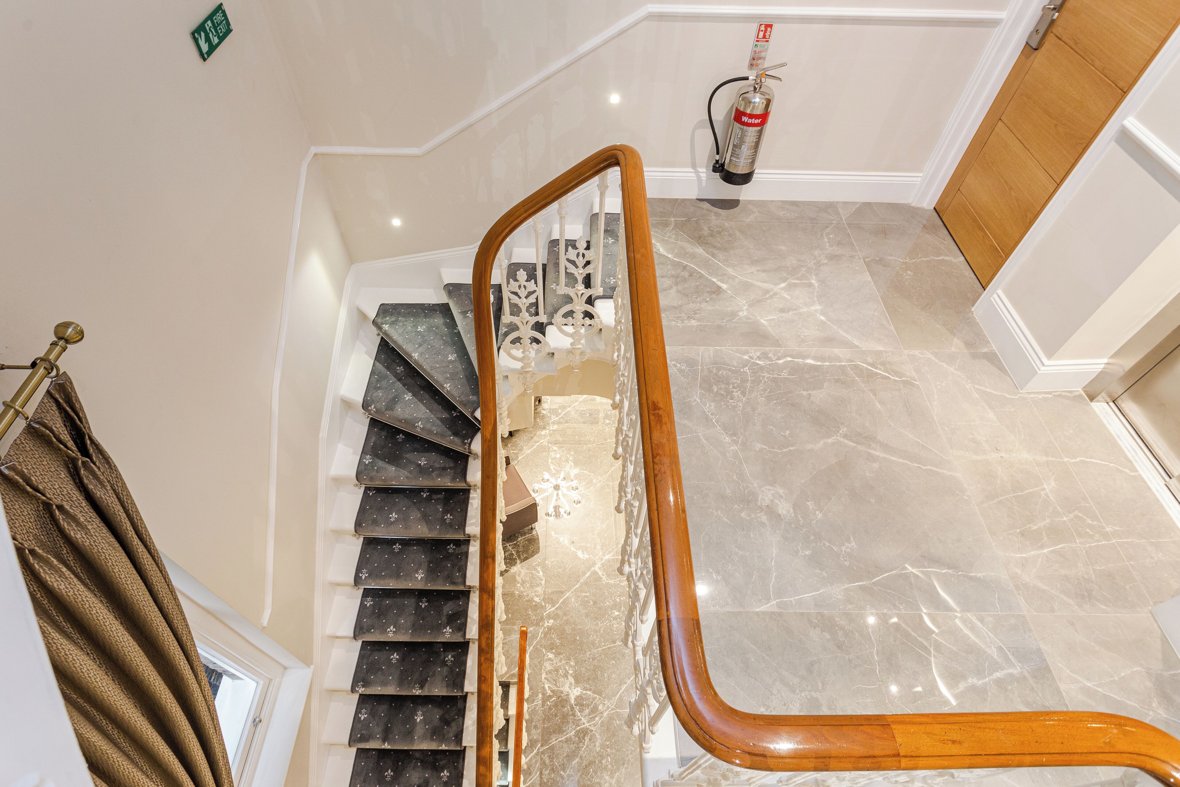 A marble staircase with a wooden handrail, a fire extinguisher mounted on the wall, and a green exit sign.
