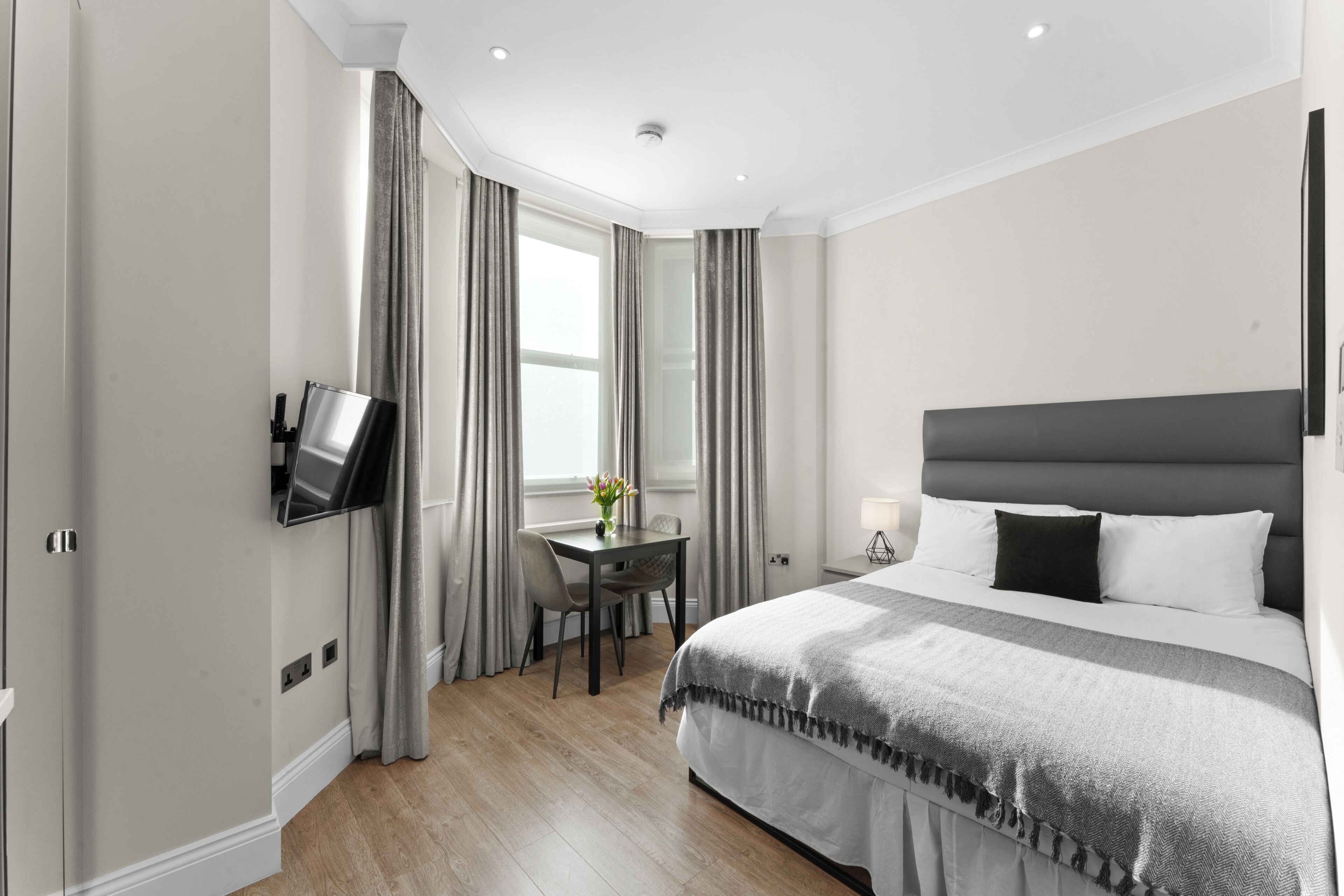 A modern hotel room with a large bed, a flat-screen TV mounted on the wall, a small table with chairs, and a window with curtains.