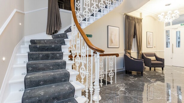 A grand staircase with ornate white balustrades and dark carpeted steps, leading to a lobby area with a chandelier and seating. - Cleveland Residences Chelsea (London)