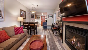 Un salon avec une cheminée, un canapé, une table à manger avec des chaises, et un coin cuisine à l’arrière-plan.