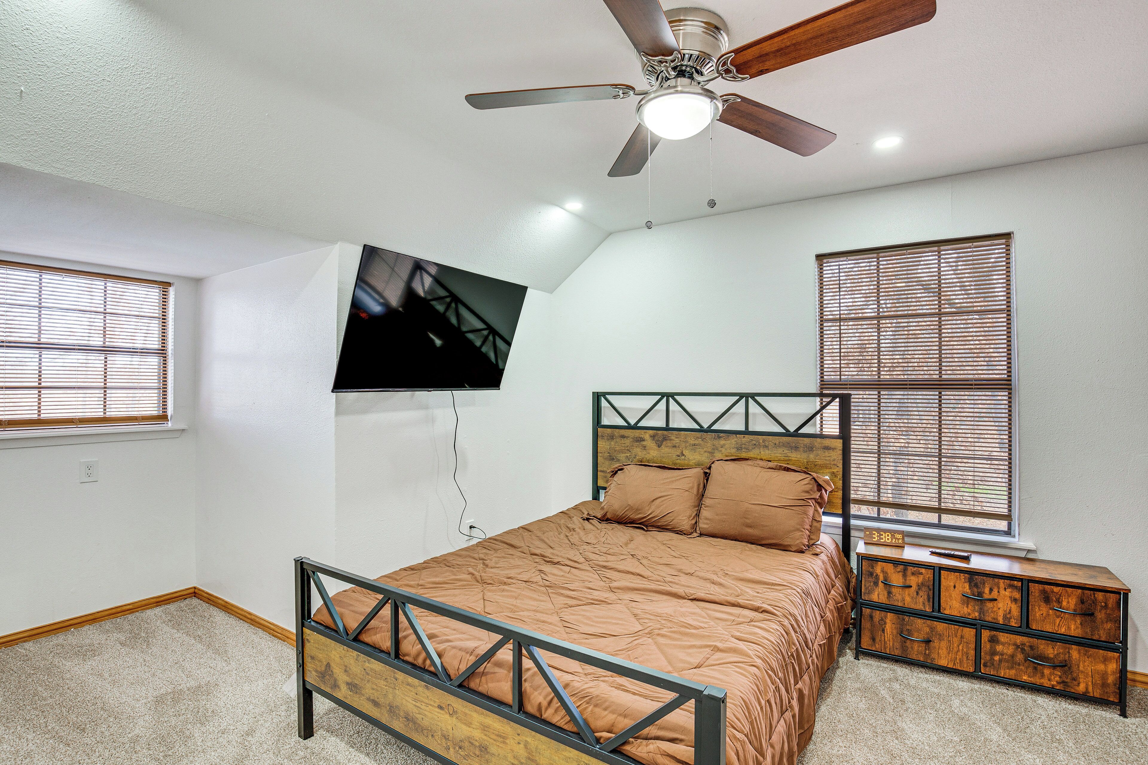 Un dormitorio con cama, ventilador de techo, televisor montado en la pared y ventana con persianas.
