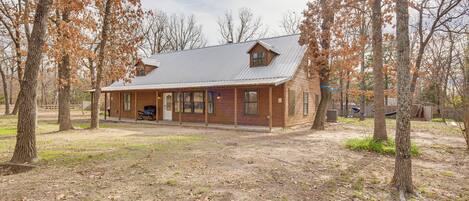 A rustic log cabin with a porch, surrounded by trees and a dirt yard.