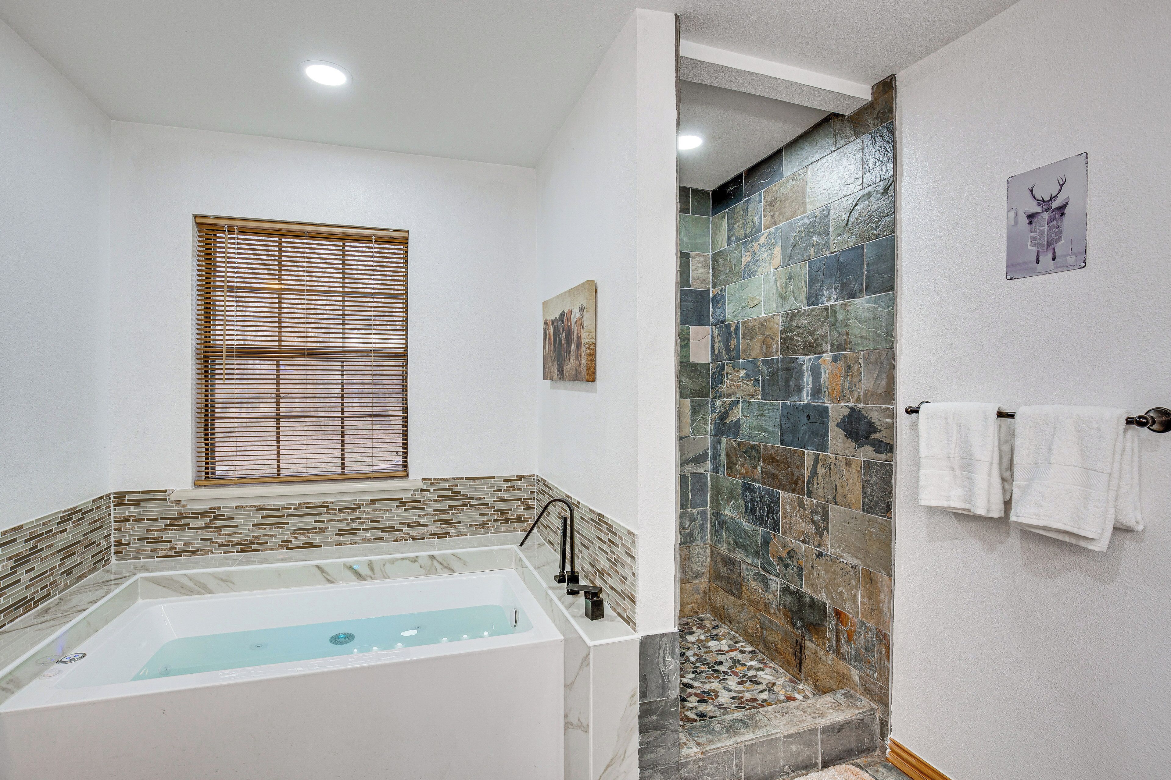 A bathroom with a bathtub, a shower with stone tiles, a window with blinds, and a towel rack.