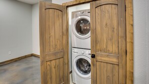 A laundry room with two stacked front-loading washing machines, wooden doors, and a concrete floor. - Peaceful Yantis Retreat - 1 Mi to Lake Fork! (Yantis)
