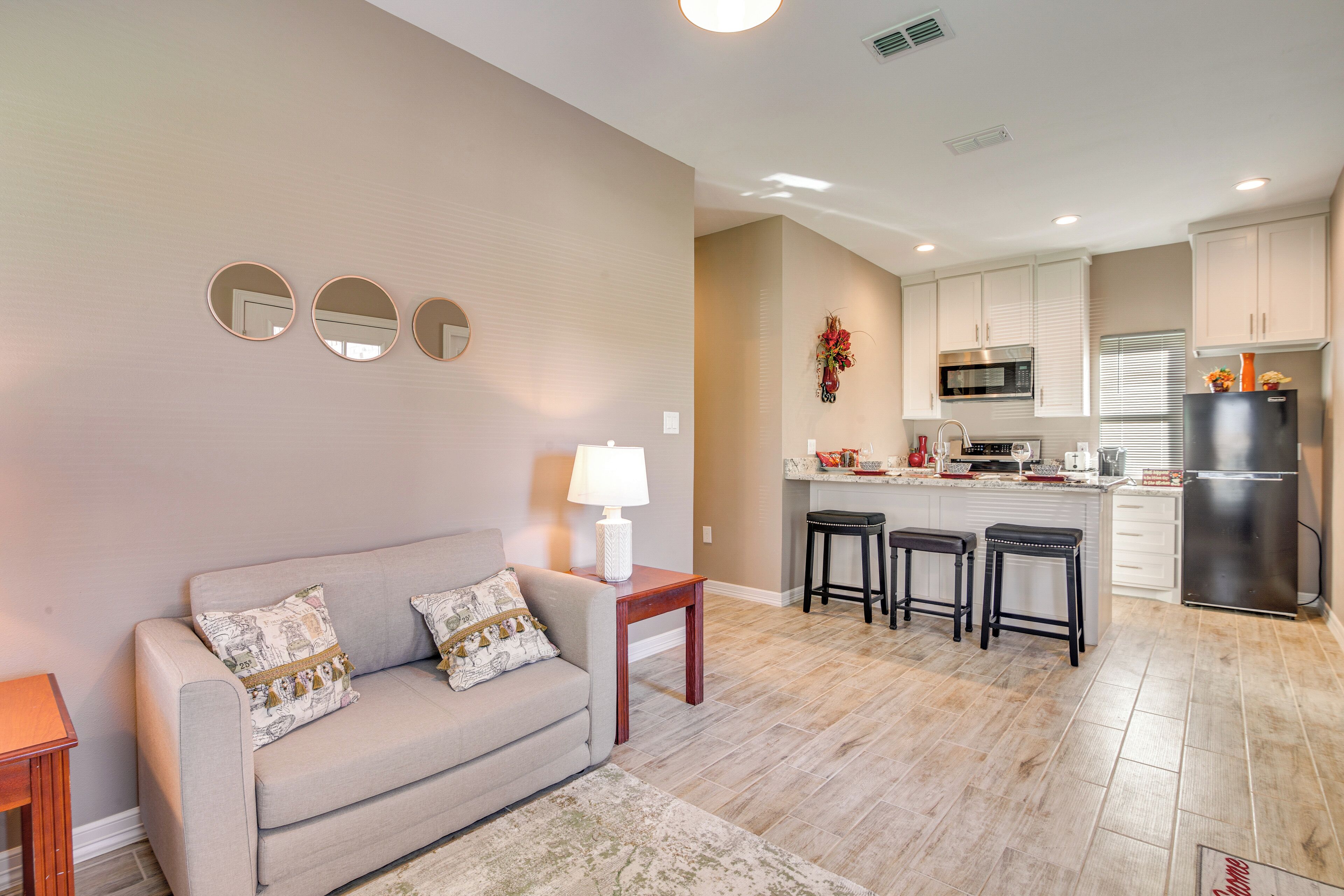 A modern living room with a sofa, a coffee table, and a rug. Adjacent to the living room is a kitchen with a microwave, refrigerator, and bar stools.
