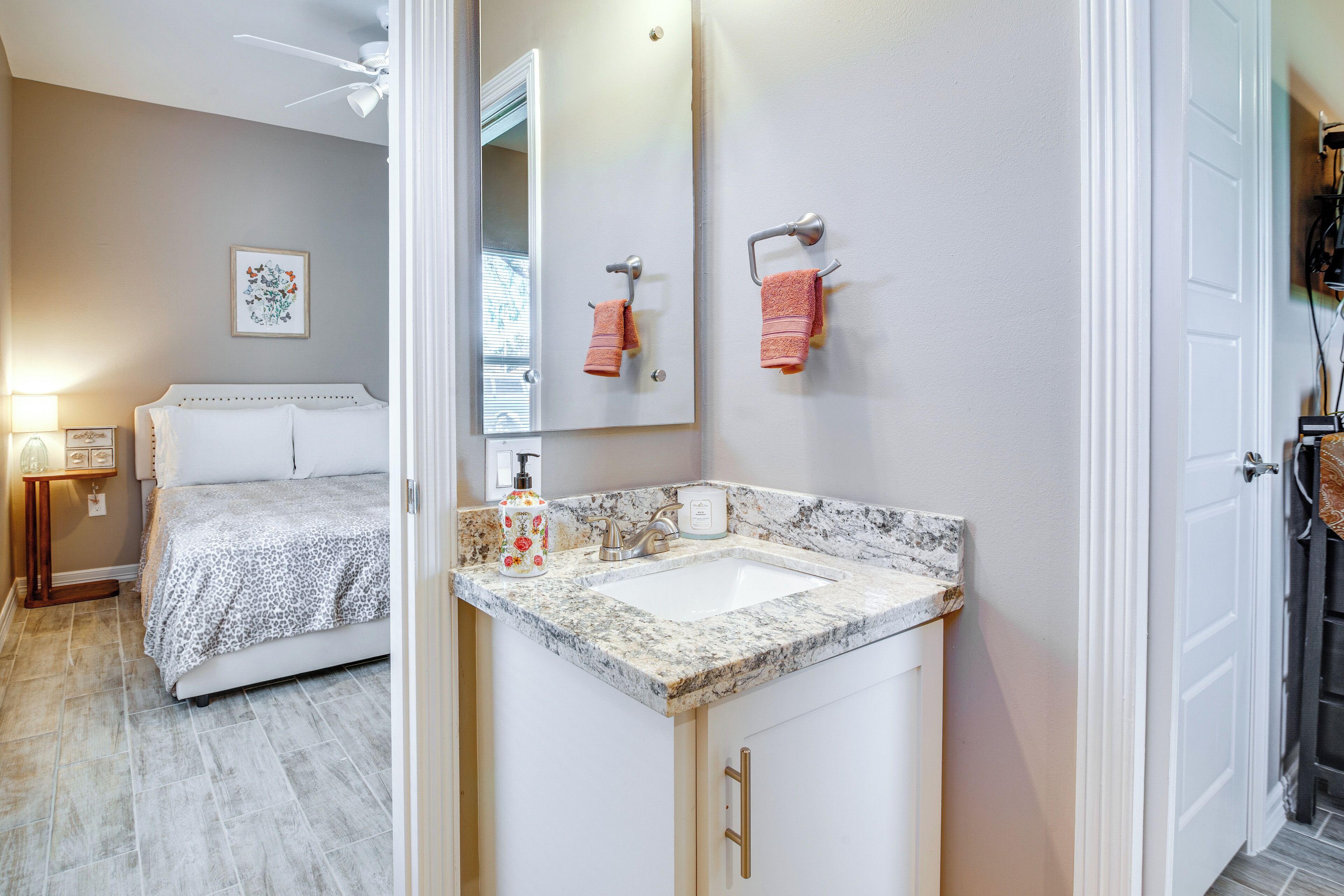 A bedroom with a bed, a nightstand, a lamp, and a bathroom with a sink, mirror, and towel rack.