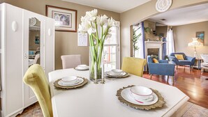 Un coin repas avec une table blanche dressée pour quatre personnes, un vase rempli de fleurs et une horloge au mur.