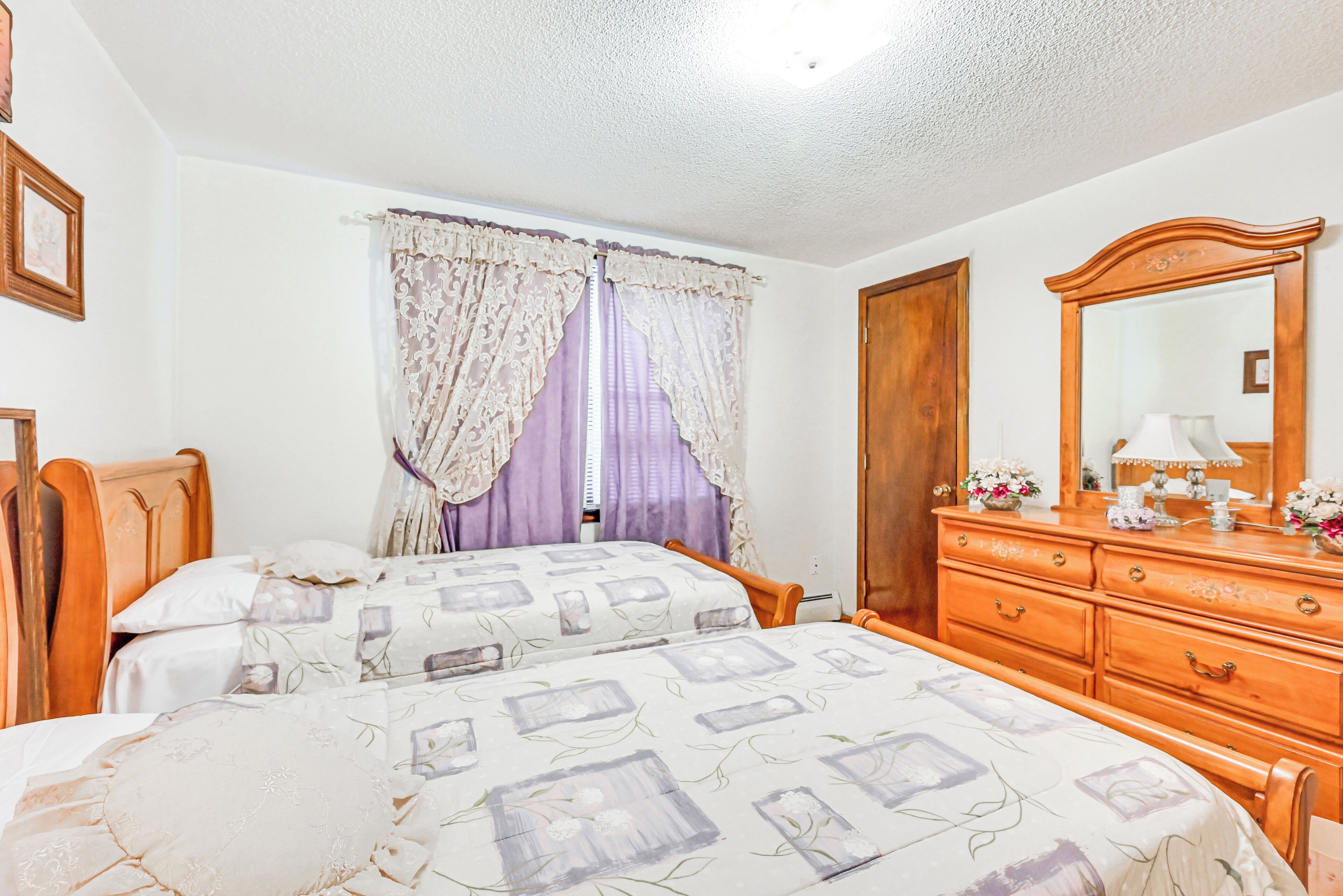 A bedroom with two beds, a wooden dresser, a mirror, and a window with purple curtains.