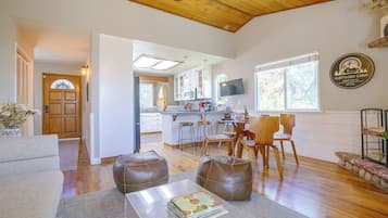 Un interior moderno con techo de madera, una cocina con armarios blancos, un comedor con sillas de madera y una sala de estar con sofá y otomanas.