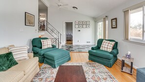 Ein geräumiges Wohnzimmer mit einem beigen Sofa, zwei grünen Sesseln, einem Holztisch und einem gemusterten Teppich. Eine Treppe mit Metallgeländer führt auf eine obere Ebene.