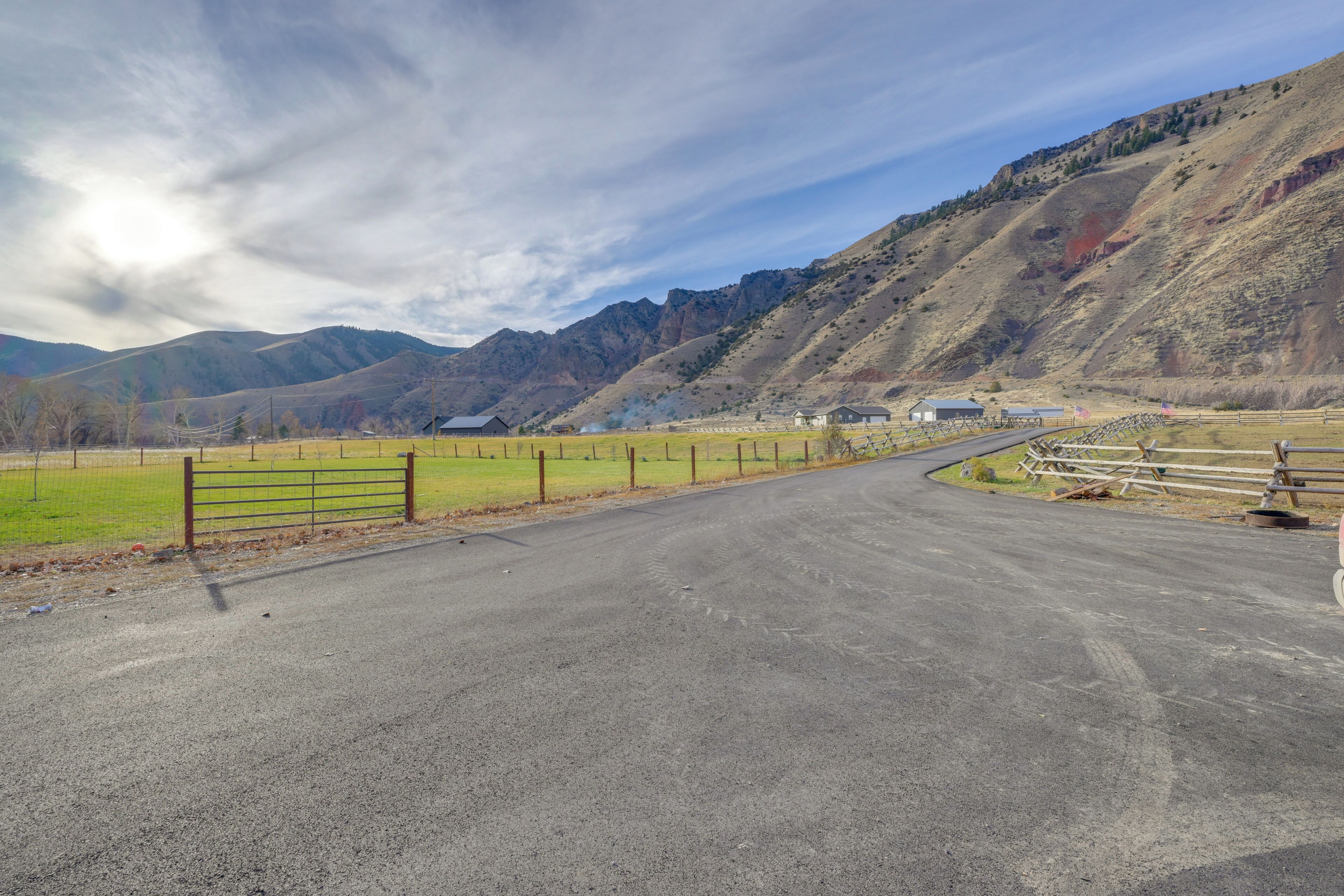 A rural road curving through a mountainous area with a fenced field and a few buildings in the distance.