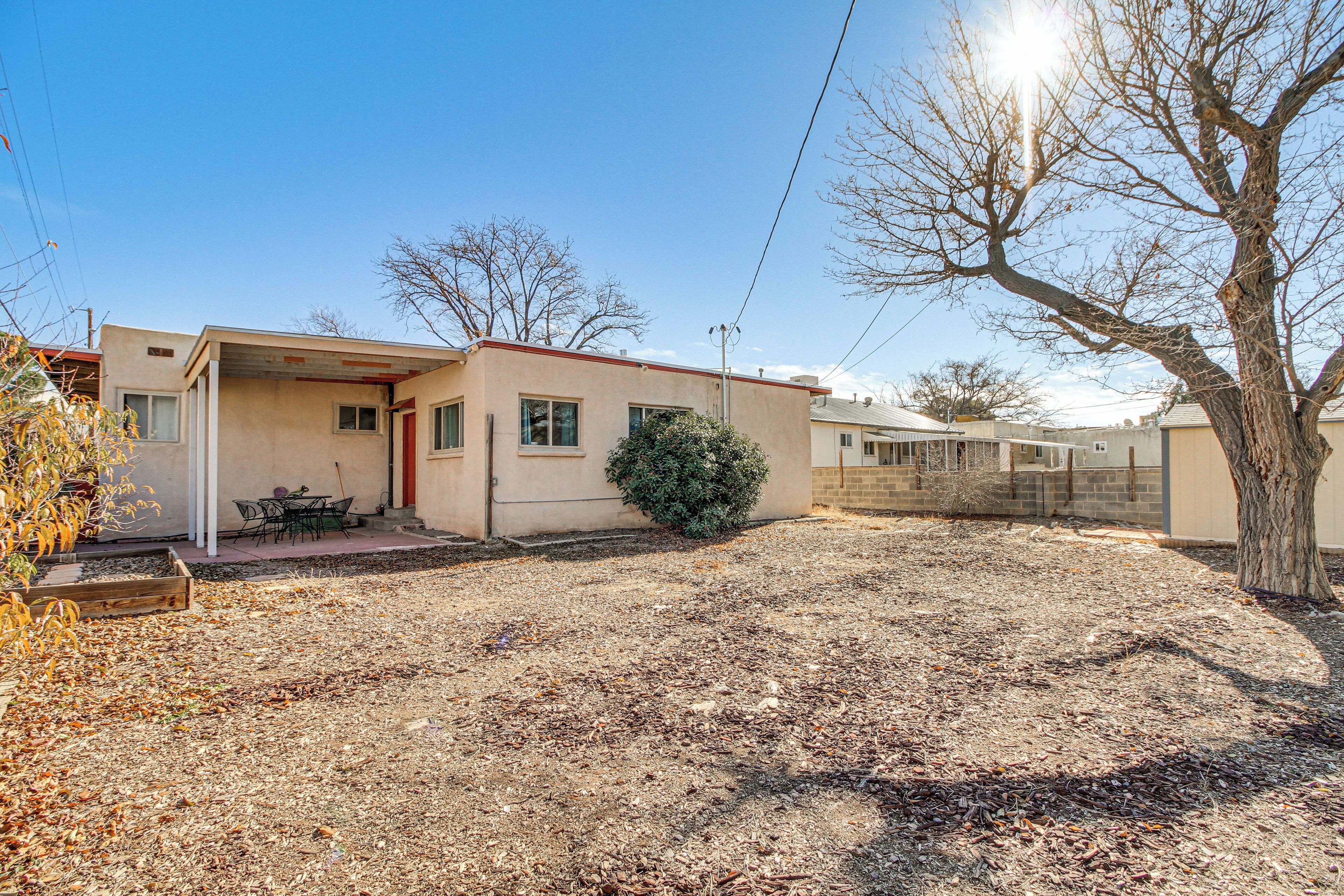 A single-story house with a small porch, a covered outdoor seating area, and a tree in the yard.