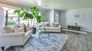 Un salon avec un canapé blanc, une table basse et une grande plante en pot. Une fenêtre donne sur l’extérieur, et une porte se trouve au fond de la pièce.