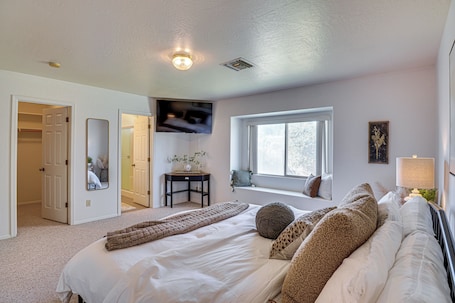 A bedroom with a large bed, a TV mounted on the wall, a window, and a bedside table with a lamp.