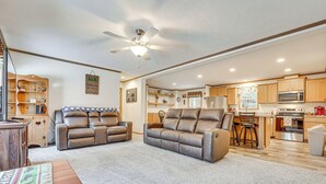 A spacious living room with a large gray sectional sofa, a wooden coffee table, and a ceiling fan. Adjacent to the living room is a kitchen with wooden cabinets, a microwave, and a refrigerator. - < 1 Mi to Beach: Peaceful Brant Lake Home! (Brant Lake)