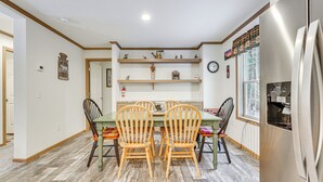 A dining area with a wooden table and chairs, a kitchen with a refrigerator, and a window with a view of trees. - < 1 Mi to Beach: Peaceful Brant Lake Home! (Brant Lake)