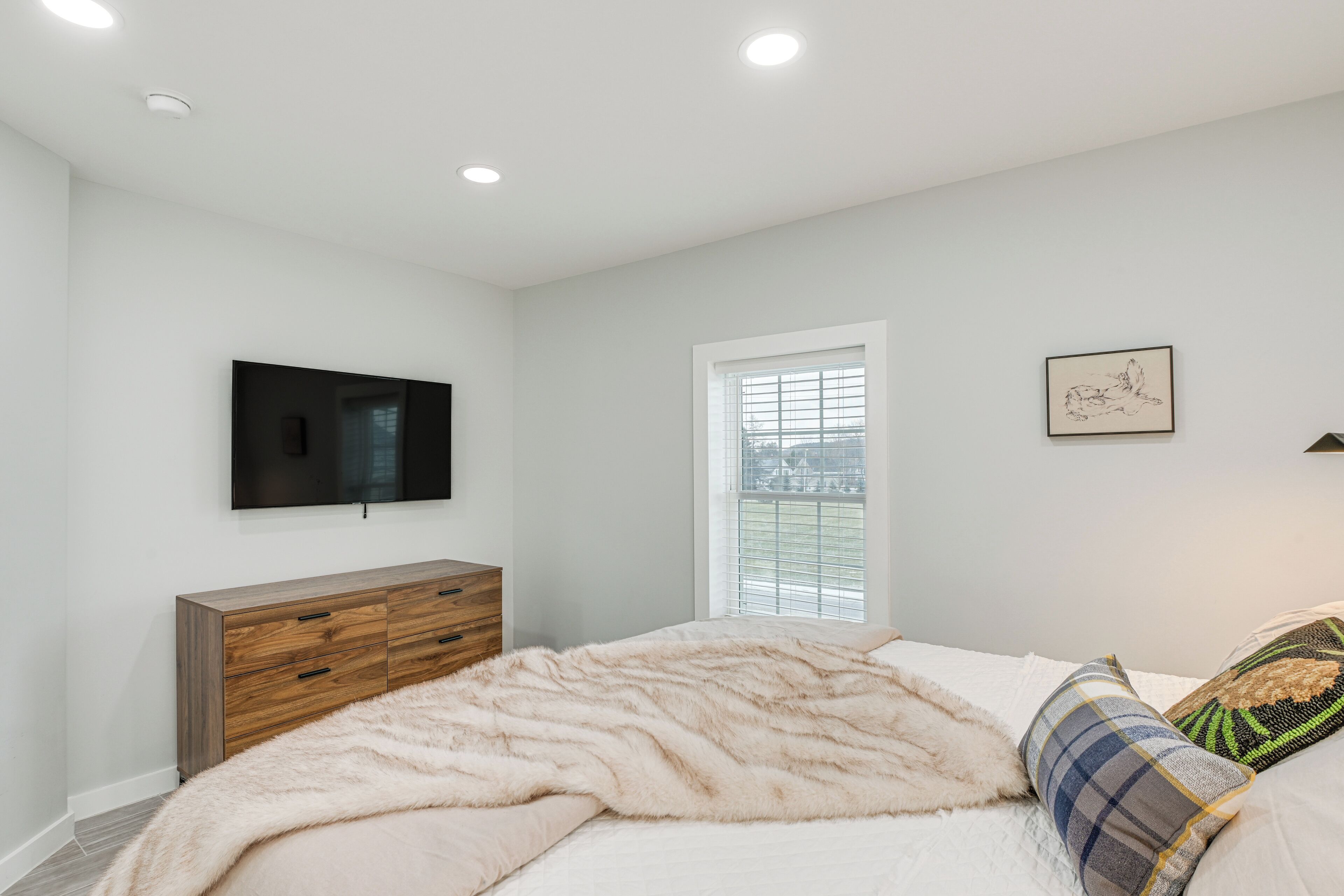 Une chambre à coucher comprenant un lit, une commode en bois, un téléviseur à écran plat fixé au mur, une fenêtre avec des stores et un tableau encadré accroché au mur.