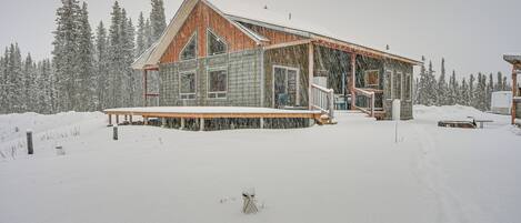A log cabin with a stone foundation and wooden exterior, surrounded by a snowy landscape and dense forest.