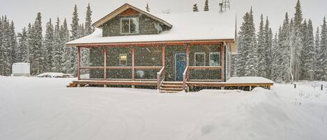 Un chalet enneigé, avec une façade en bois, de grandes fenêtres et un porche à l’avant.