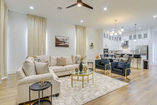 A modern living room with a sofa, coffee table, and chairs, a kitchen with cabinets and appliances, and a ceiling fan.