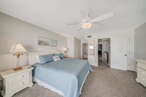 Ein Schlafzimmer mit Bett, Nachttischlampen, Deckenventilator, einem Gemälde und einem Flur, der zu einem Badezimmer führt.