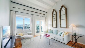 Un soggiorno moderno con un divano bianco, un tavolino in vetro e una TV su un supporto in legno. Sono presenti ampie finestre con tende, vista sulla città e due specchi fissati alla parete.