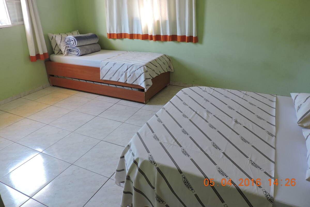 A room with two beds, one larger and one smaller, both with striped bedding. There is a window with curtains, a tiled floor, and a green wall.