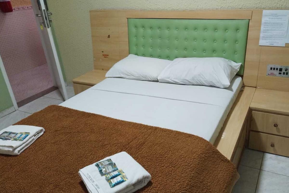 A single bed with a green upholstered headboard, white bedding, and a brown throw blanket. There are two bedside tables with drawers, one of which has a paper attached.