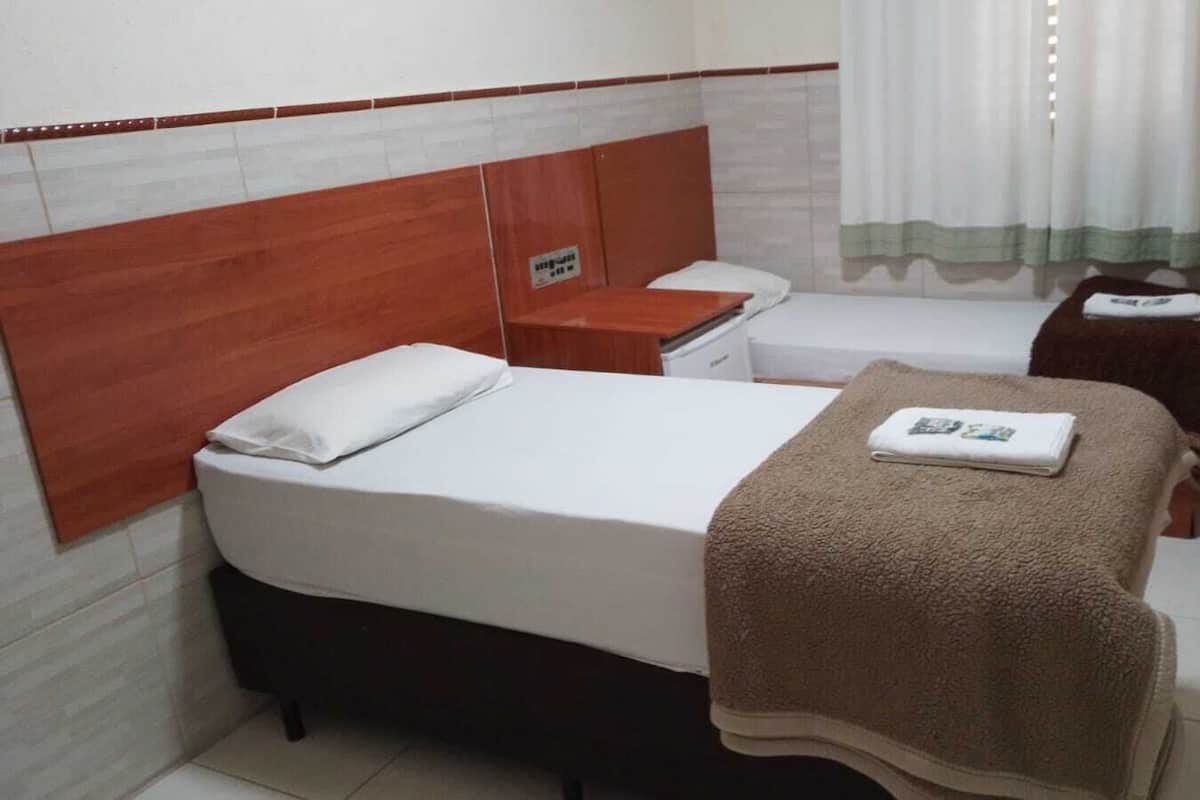 A hotel room with a bed, a bedside table, a small table, a chair, and a window with curtains.