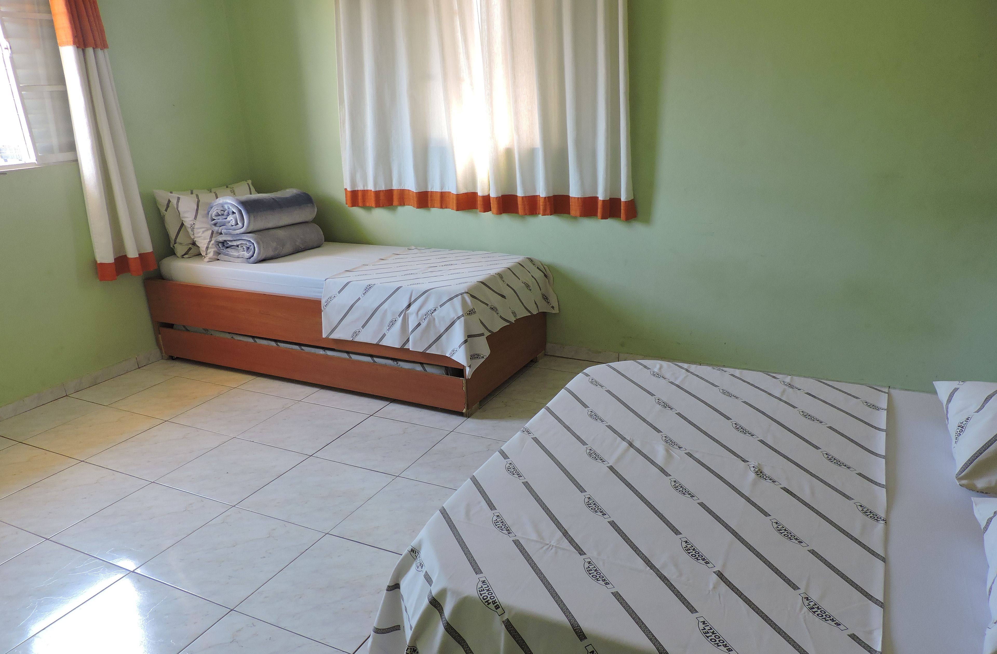 A room with two beds, one larger and one smaller, both with striped bedding. There is a window with curtains, a tiled floor, and a green wall.