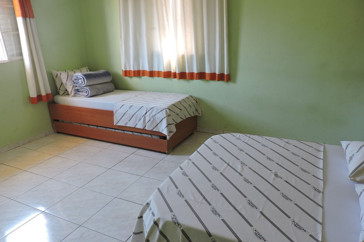 A room with two beds, one larger and one smaller, both with striped bedding. There is a window with curtains, a tiled floor, and a green wall.