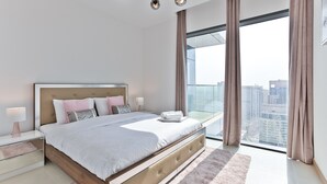 Un dormitorio moderno con una cama grande, lámparas de noche, vistas de la ciudad a través de ventanas del suelo al techo y un paisaje urbano visible desde el exterior.