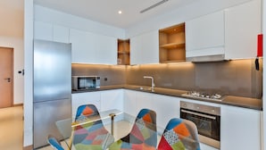 Una cocina moderna con armarios blancos, un refrigerador de acero inoxidable, un microondas, una estufa a gas y una mesa de comedor de vidrio con sillas de colores.