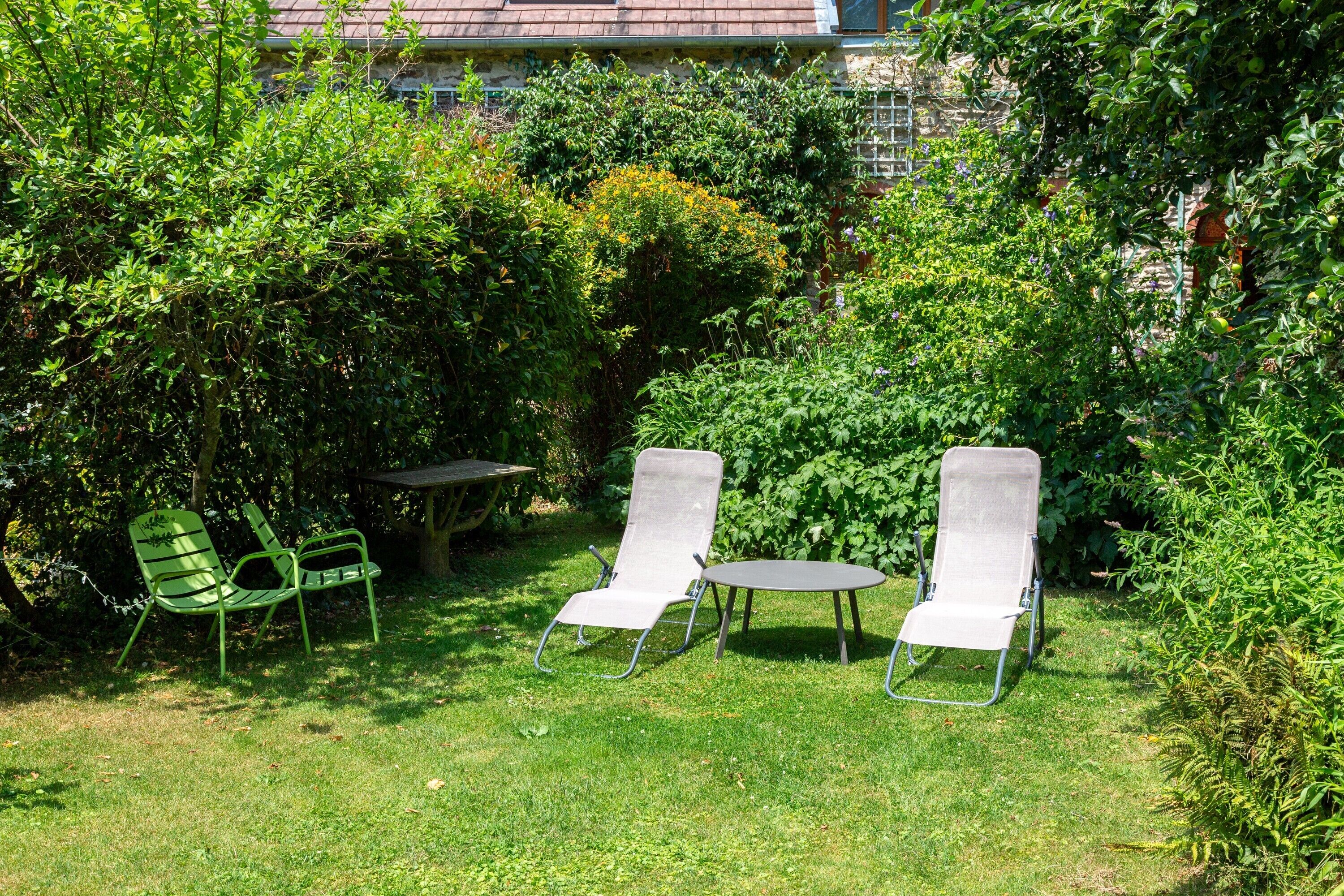 Un jardin avec deux chaises en métal et une petite table, entouré d’un feuillage dense et avec un bâtiment visible au loin.