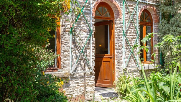 A stone building with arched windows and a wooden door, surrounded by greenery.