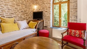 A room with a stone wall, a television, a red chair, a wooden table, and a window with a view of greenery.