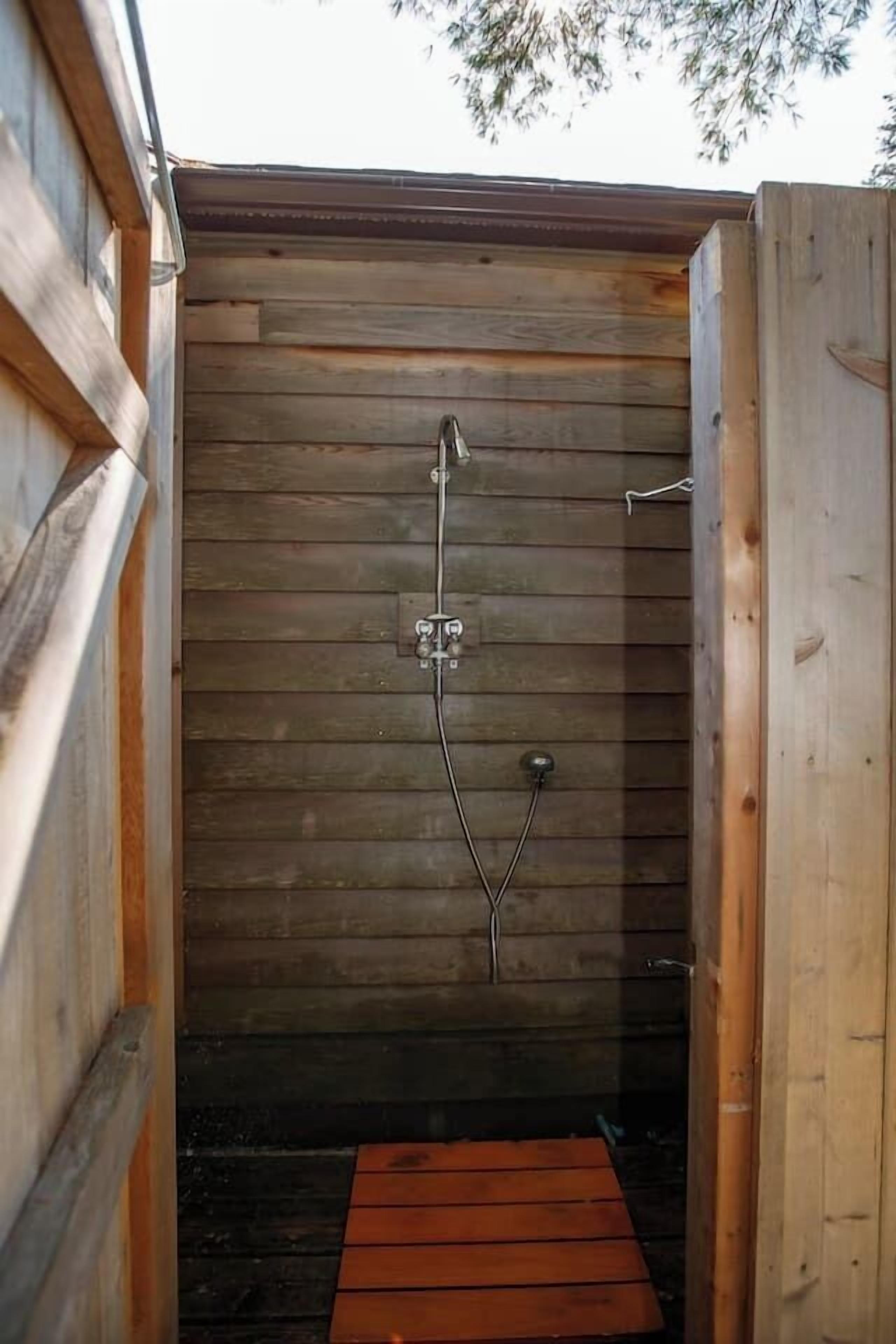 Een houten douchecabine met een metalen douchekop en een klein houten treetje om erin te stappen.
