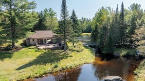 Une cabane au bord d’une rivière, avec une terrasse et un petit pont.