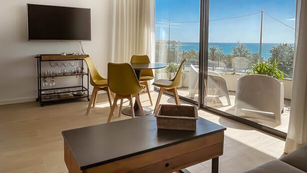 A modern living room with a large window, a flat-screen TV, a dining table with yellow chairs, and a small balcony with plants. - Modern sea Front Sea Waves (Alicante)