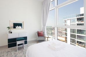 Une chambre d’hôtel moderne avec un grand lit, une petite table de chevet, une chaise, une coiffeuse avec un miroir et une vue sur la ville grâce à de grandes fenêtres.