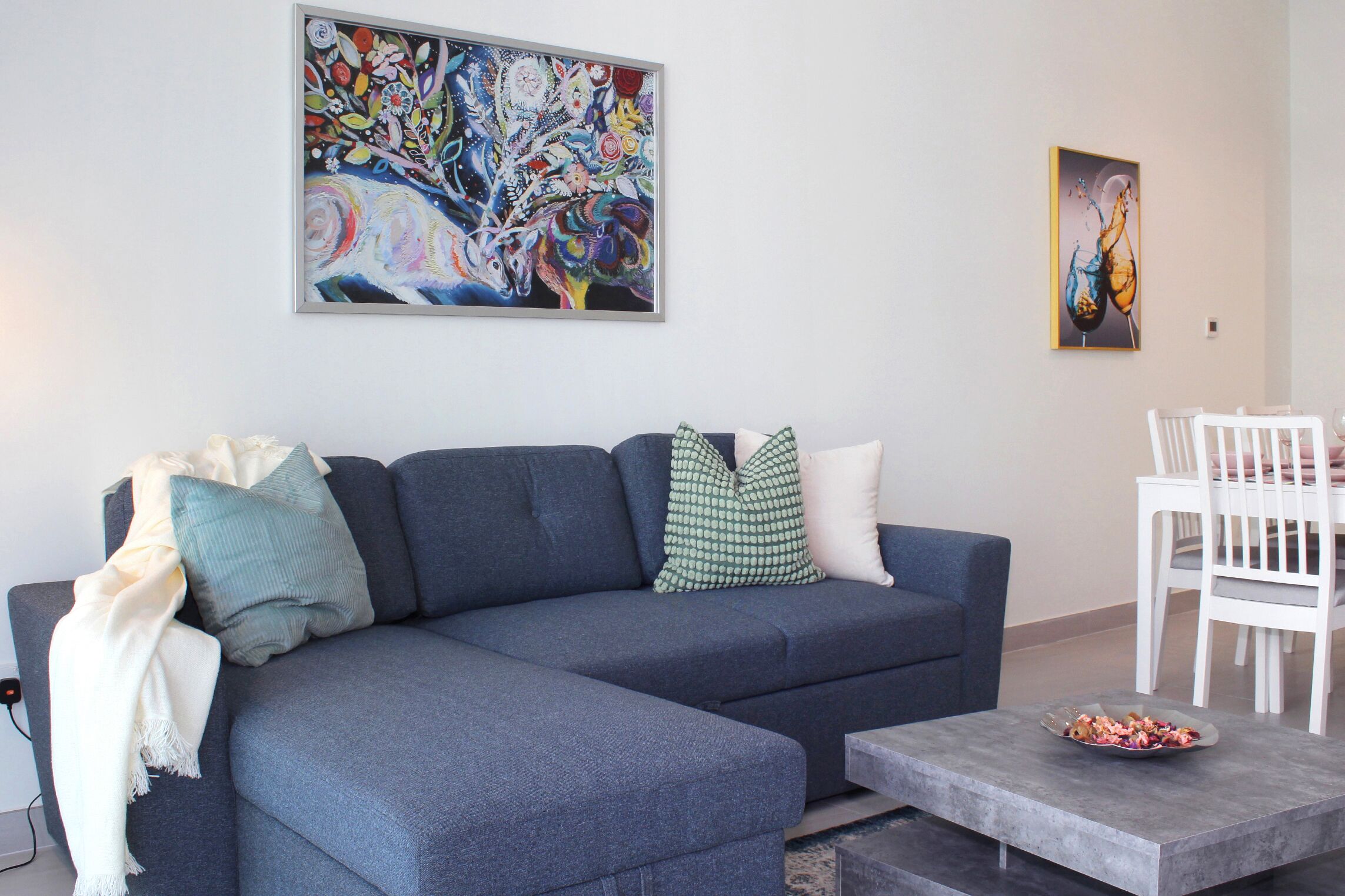 A modern living room with a grey sectional sofa, a white dining table, and a colorful abstract painting above the sofa.
